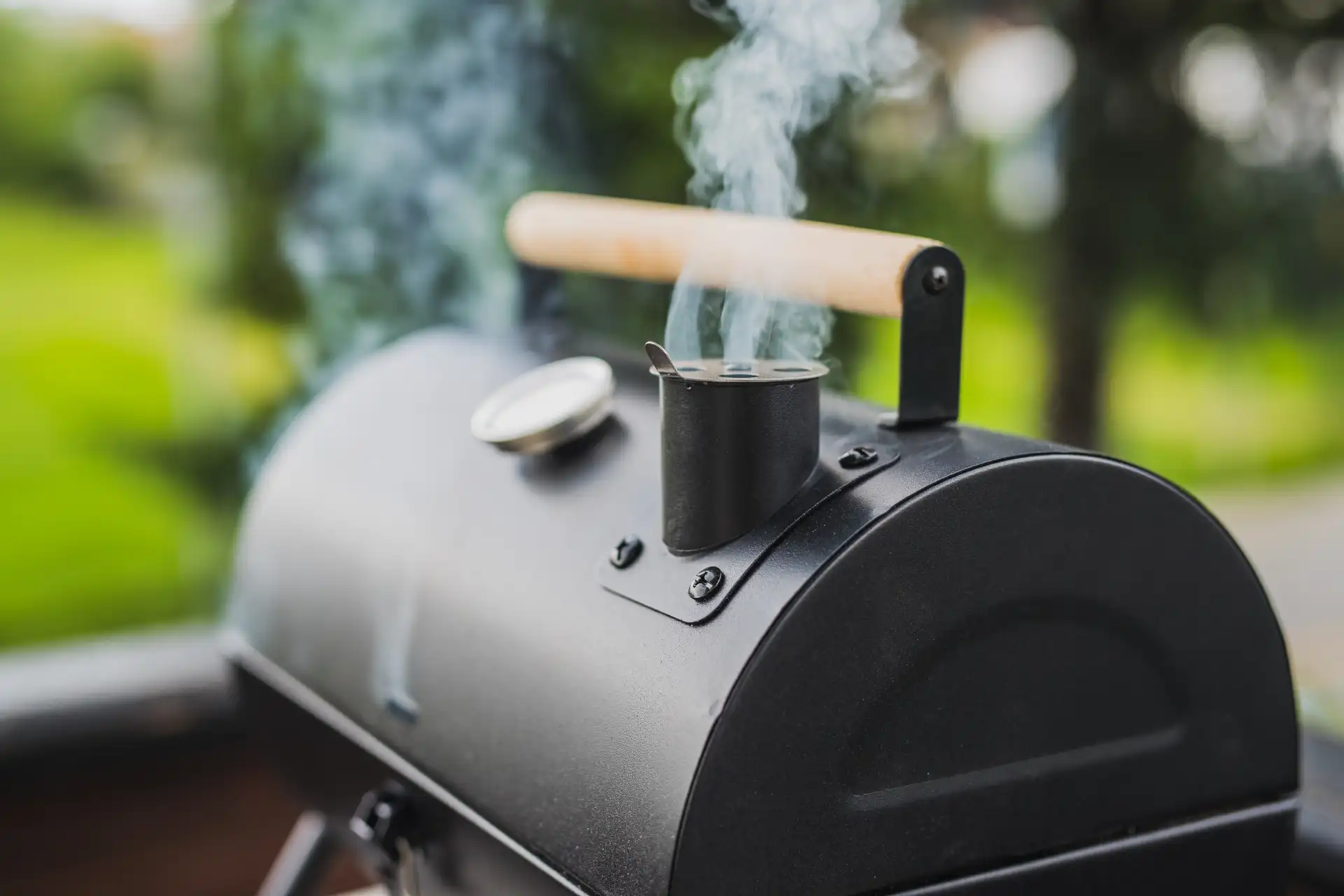 Nahaufnahme eines schwarzen Smokers mit Holzgriff, Thermometer und Rauch aus dem geöffneten Kamin